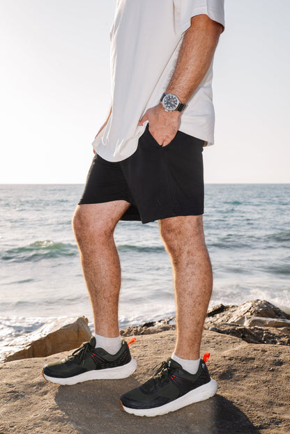 Person wearing black shorts and black sneakers on a rocky beach with ocean in the background