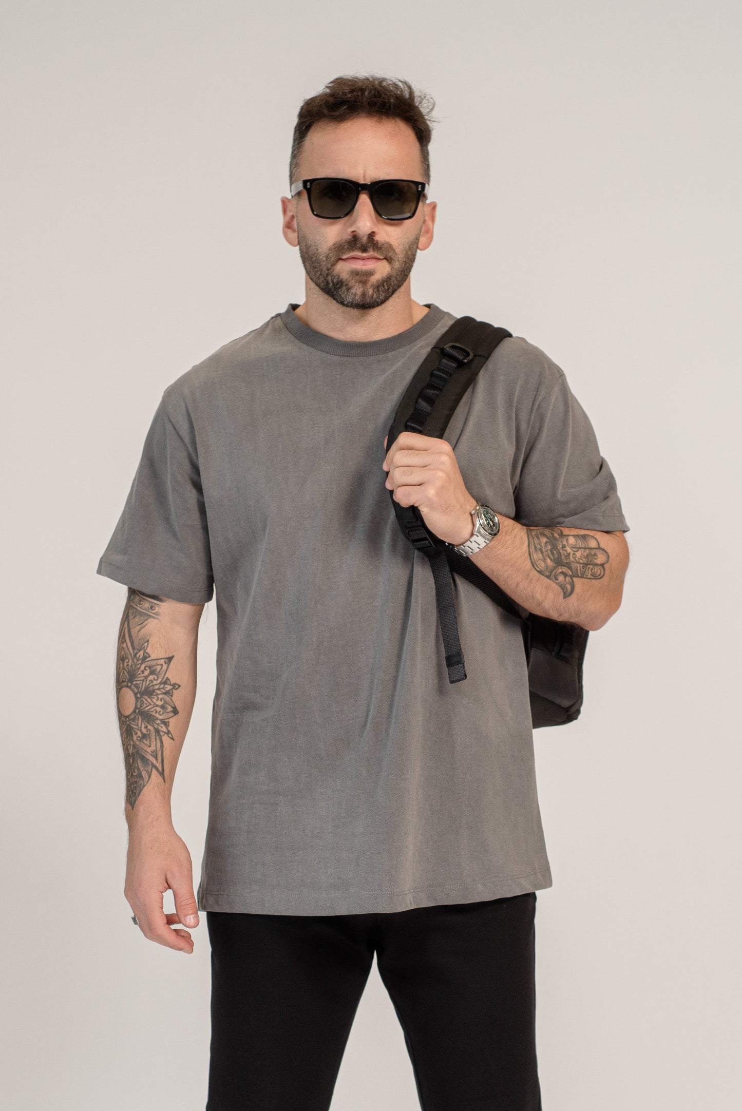Man wearing a gray t-shirt, black pants, and sunglasses, holding a black bag on a white background